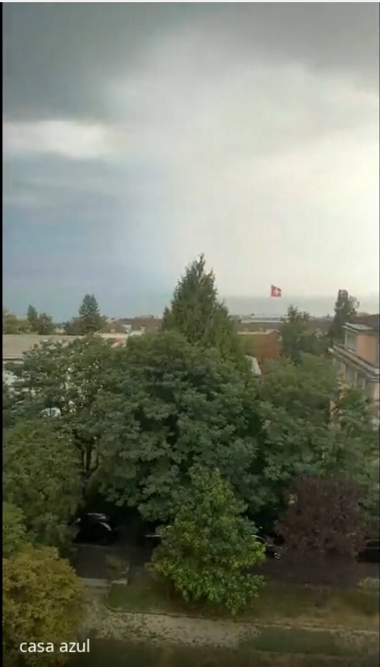 O lago Léman visto da janela de Fabrice, a tempestade se formando sobre a água, a bandeira suíça ao longe